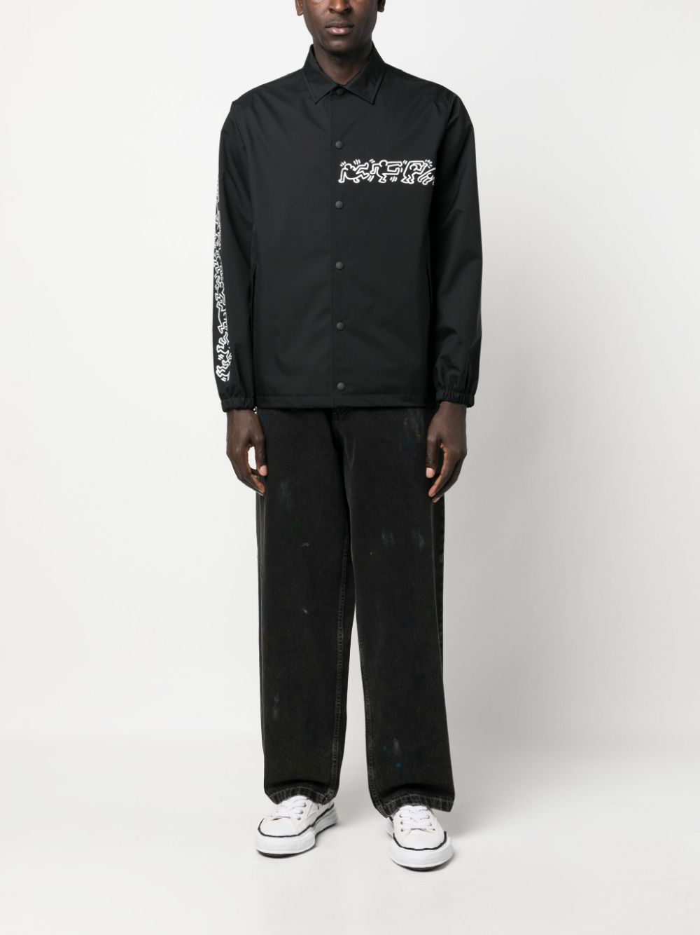 Model draagt de zwarte coach jacket bij een zwarte broek en witte sneakers