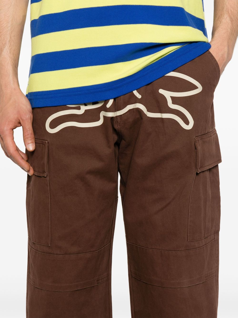 Detail van de bruine cargobroek op het model, met cargozakken met klep en de Running Dog-print