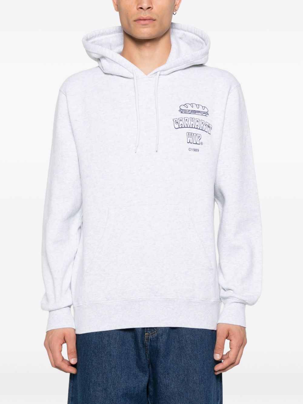 Model draagt het lichtgrijze Carhartt WIP sweatshirt met verstelbare capuchon en kangoeroezak