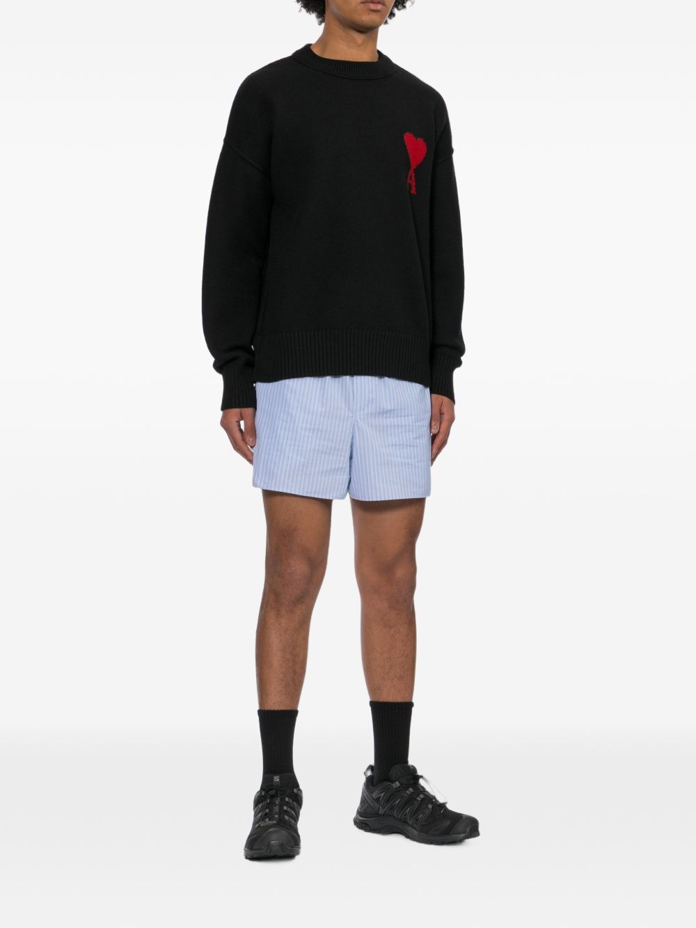Model in volledige outfit met de zwarte AMI Paris Ami de Coeur trui, gestreepte short en sneakers